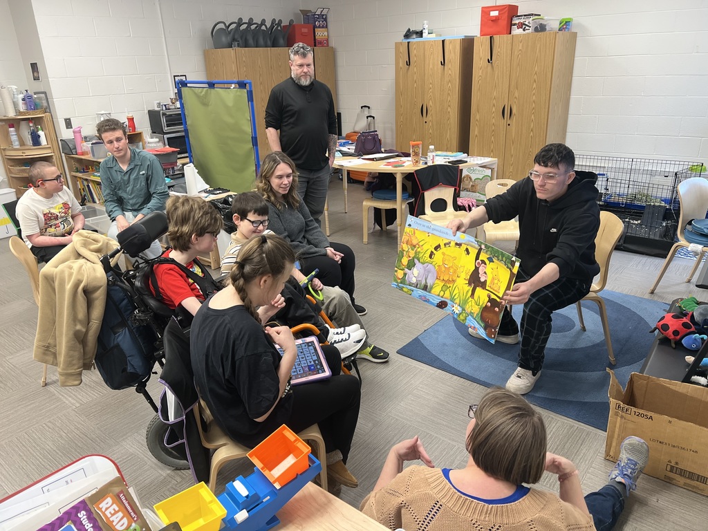 A male high school student seated in front of an elementary class holds a book open wide so the students can see the illustration. 
