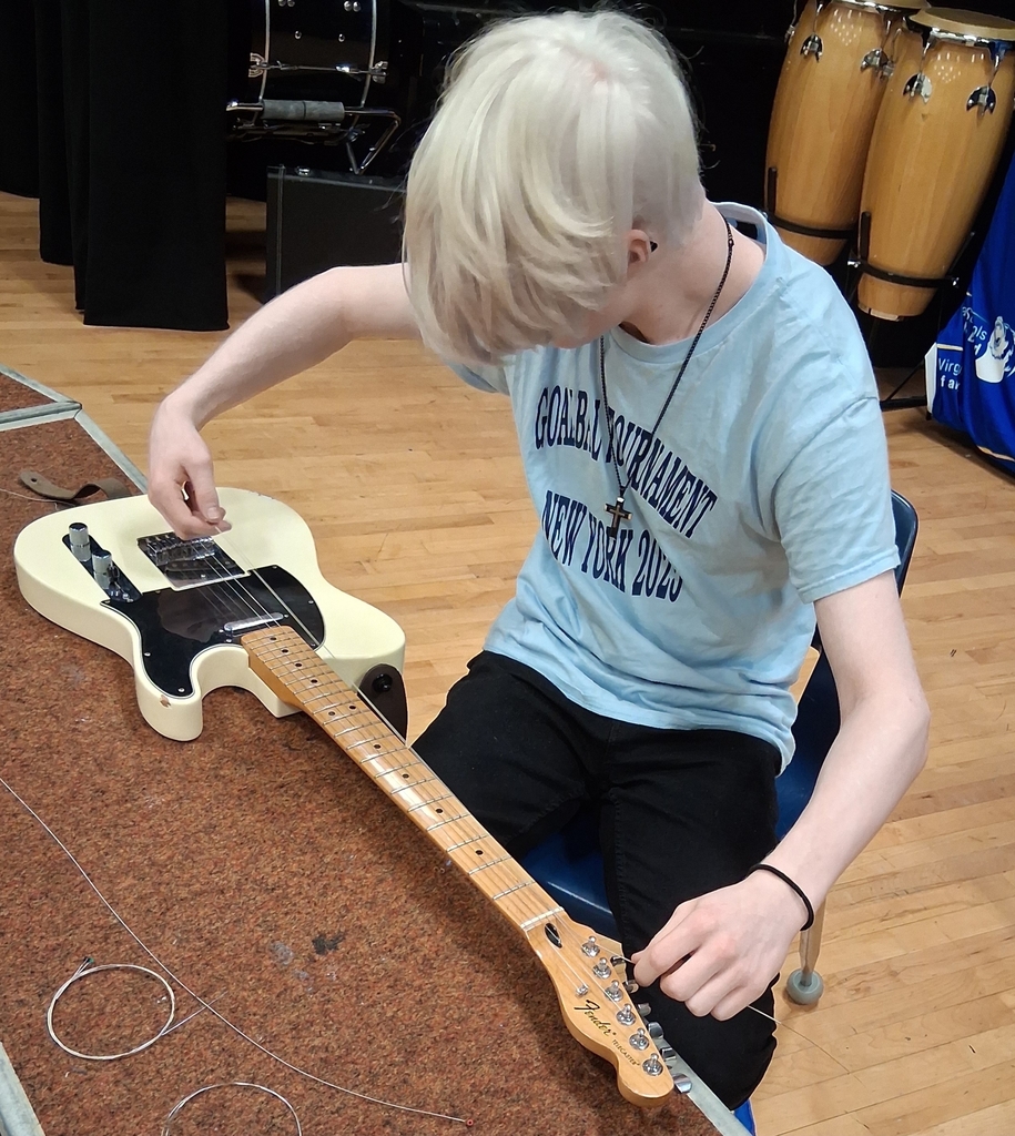 A male student is seated as he looks down and restrings a guitar. 