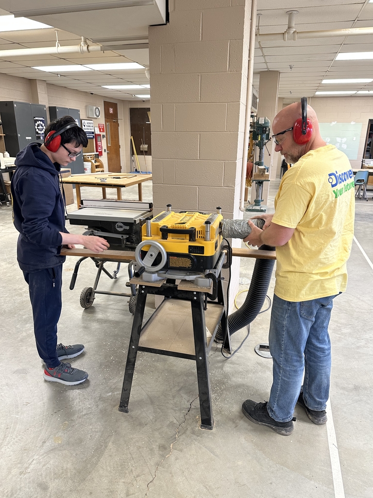 A male student and a male instructor stand on either side of a wood shop machine as the piece of wood passes through the machine to the other person. 
