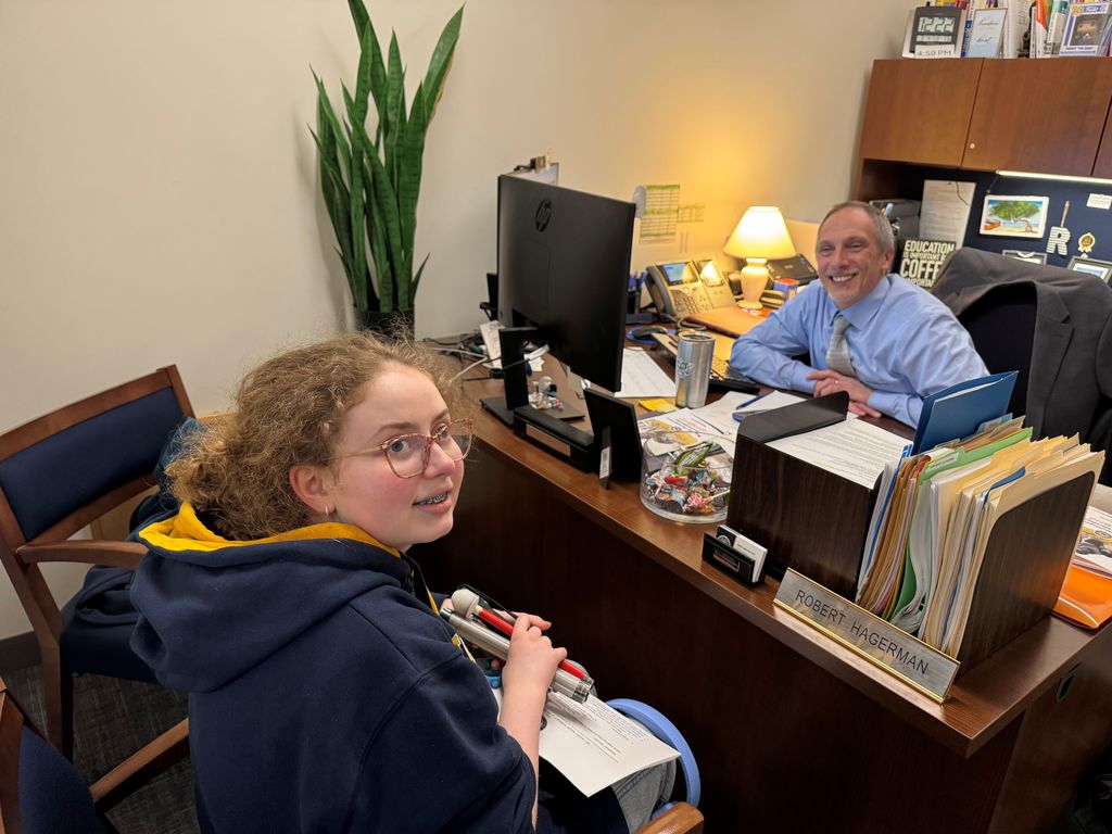 A female WVSDB student holds her cane and looks back to smile at the camera. There is a male WVDE employee sitting at a desk across from her. 