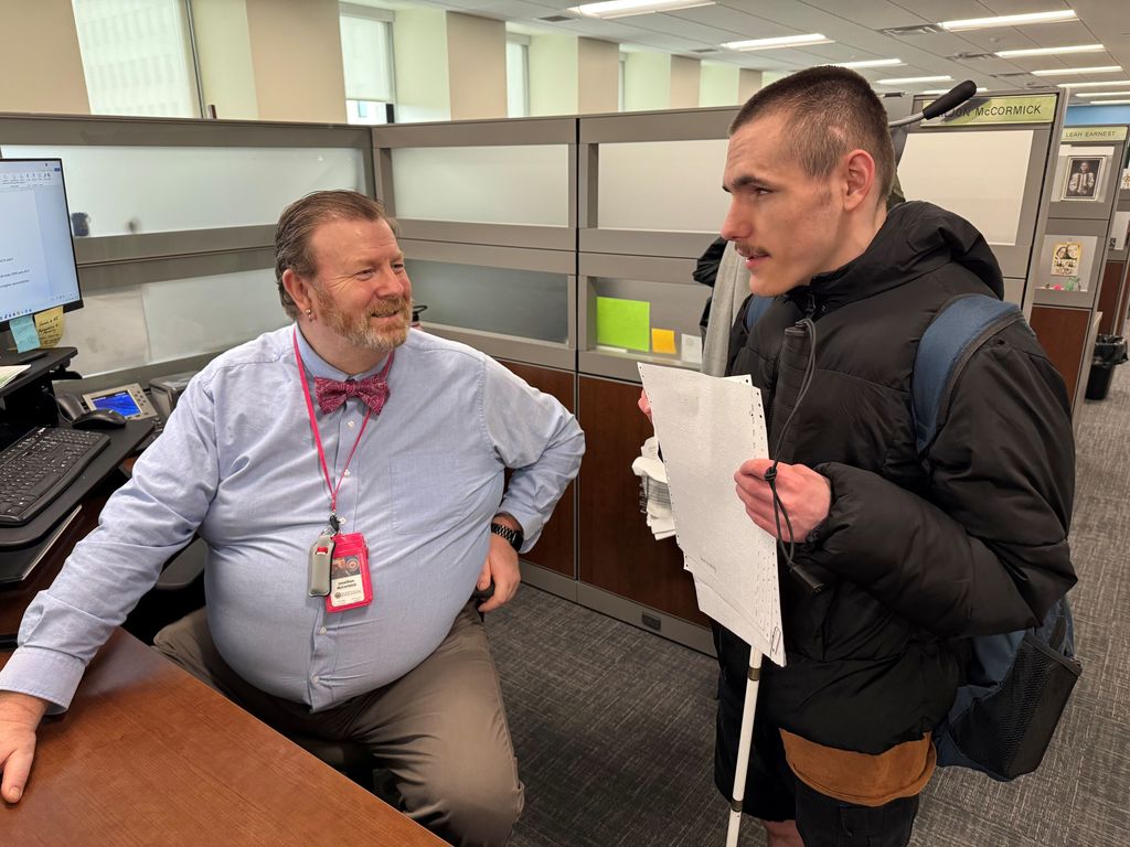 A male WVDE employee is seated in his cubicle and smiles at a male WVSDB student who is holding his cane and an embossed document in his hands. 