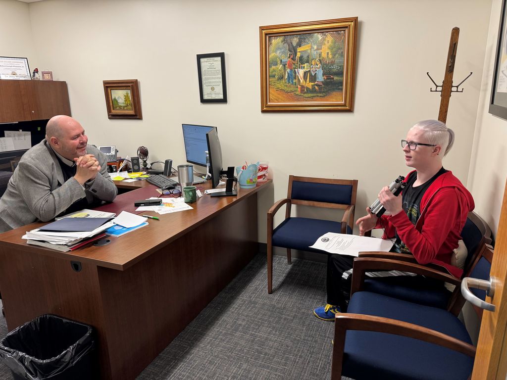 A male WVDE employee sits at his desk while a male WVSDB student is seated across from him in a chair.