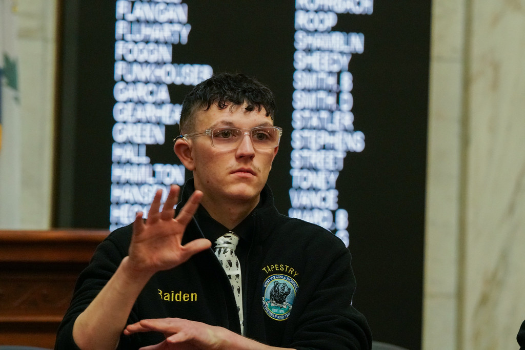 A male WVSDB student wearing a black, fleece jacket uses American Sign Language (ASL) to interpret the performance during the legislative event. 
