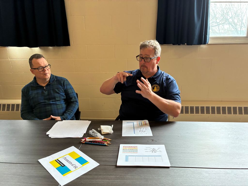 A male participant signs using American Sign Language (ASL) while a male to his right observes him.