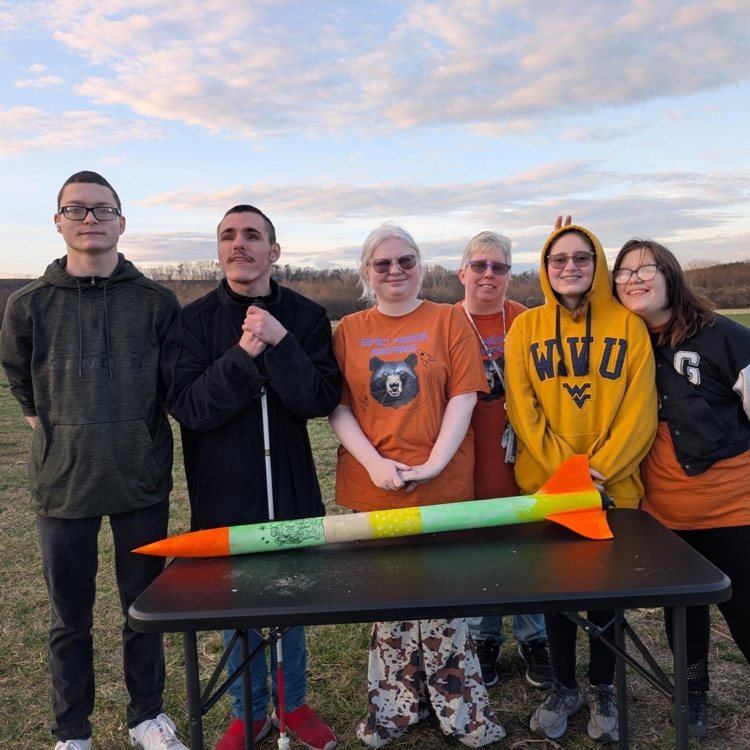 A group photo of the rocketry club with Coach Biddle. There is a green and orange rocket sitting on a table in front of the group. 