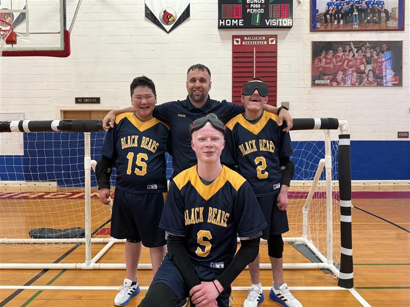 Male Goalball players and their coach, Coach Smith stand together in a group in front of a Goalball net.