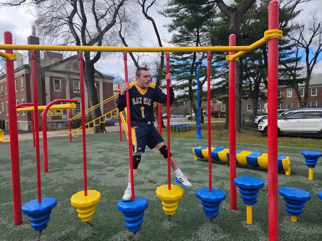 A male Goalball player dressed in a blue and gold jersey is standing on a piece of playground equipment. 
