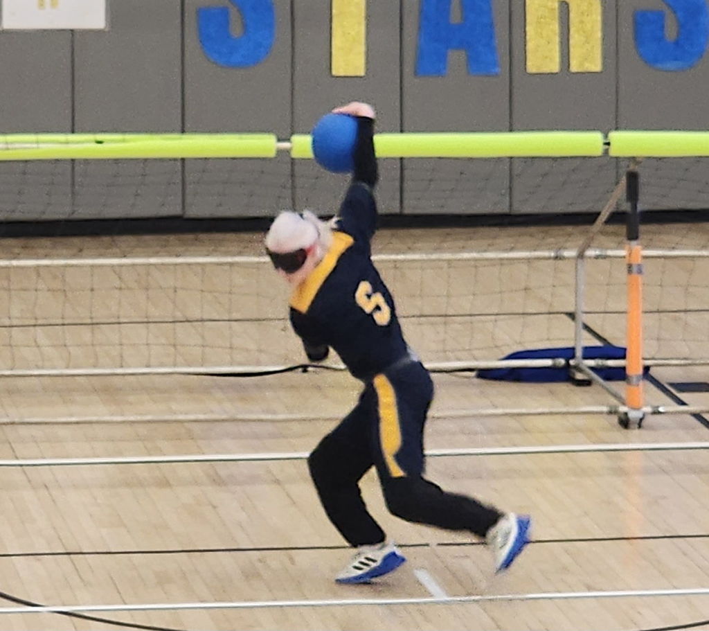 A male Goalball player wearing a blue and gold jersey gets ready to throw a blue-colored goalball.