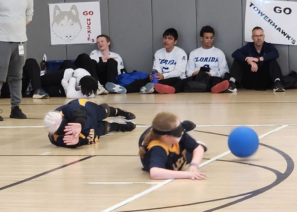 Female Goalball players wearing blue and gold jerseys laying on the floor to defend against the opposing team.