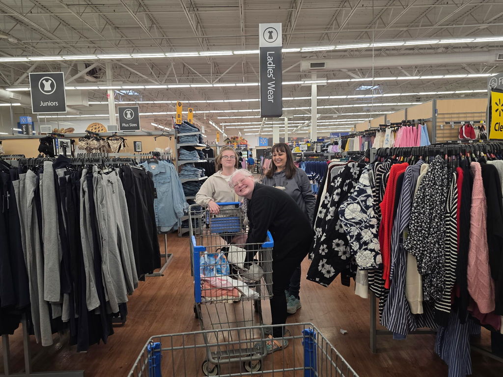 A female WVSDB representative smiles and observes two female students. One student puts items in a shopping cart while the other female student waits to push the cart.
