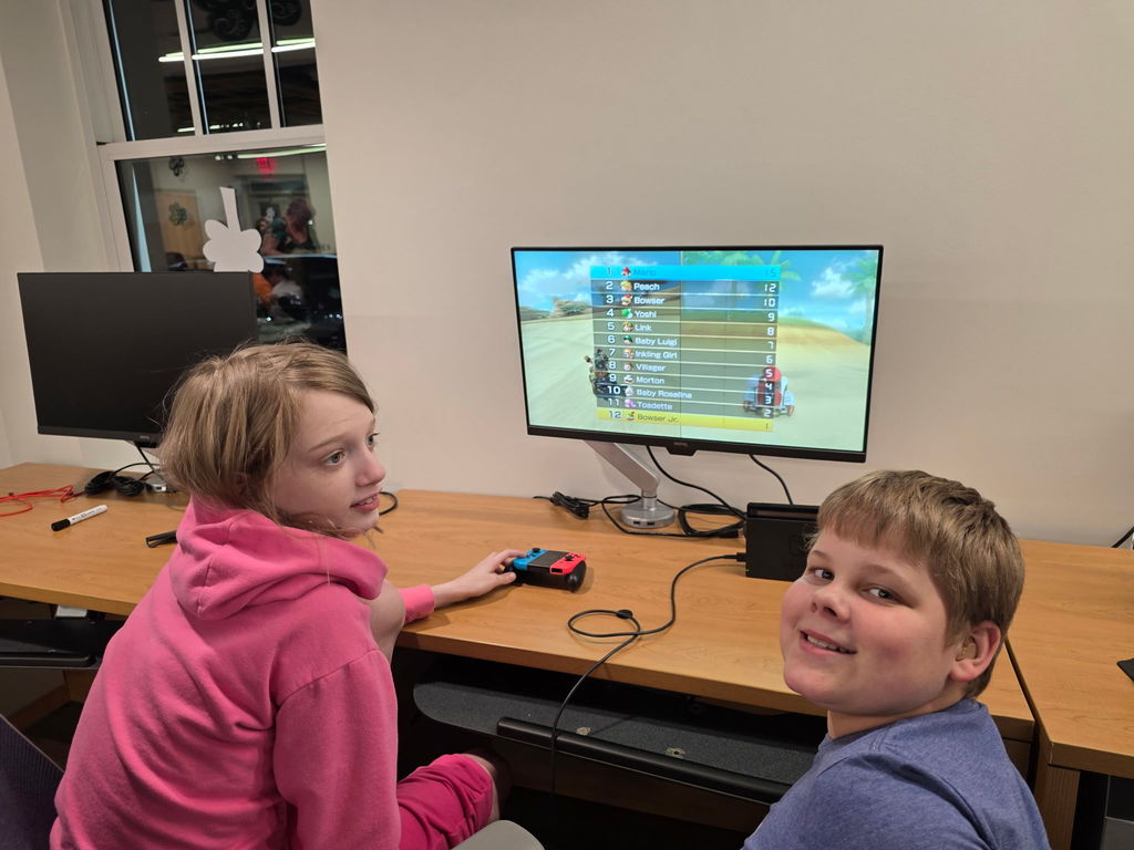 A female and a male student are seated at a gaming station. They look behind them to smile for the camera.