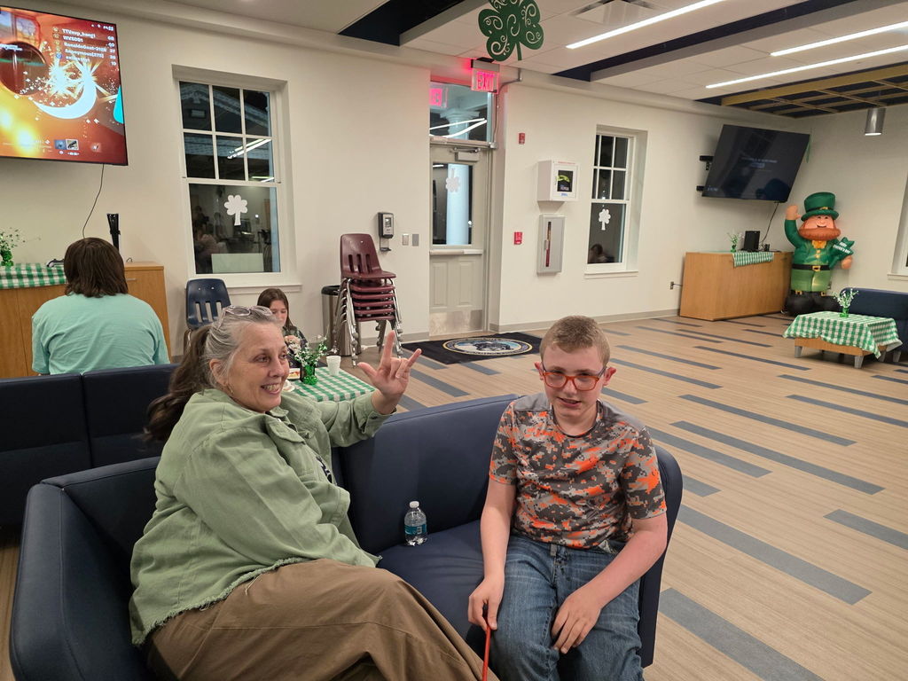 A male student and a female individual are seated on a blue-colored couch. The female flashes up the "I love You" sign in ASL.