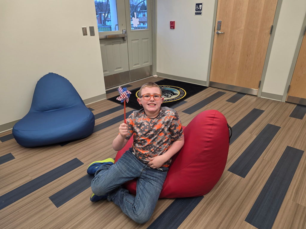 A male student sits on a red bean-bag chair and smiles for the camera.