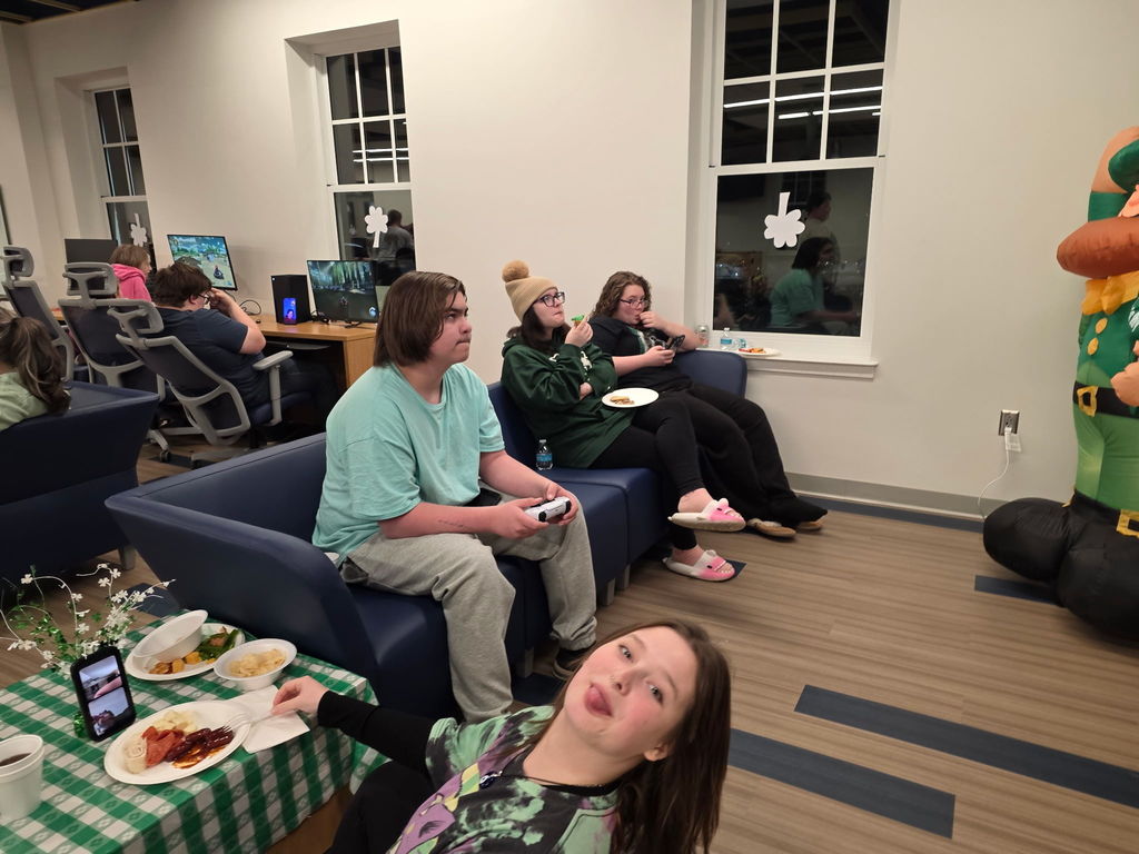 Students seated on a blue-colored couch as they eat snacks and play video games. A female in the foreground sticks out her tongue at the camera.