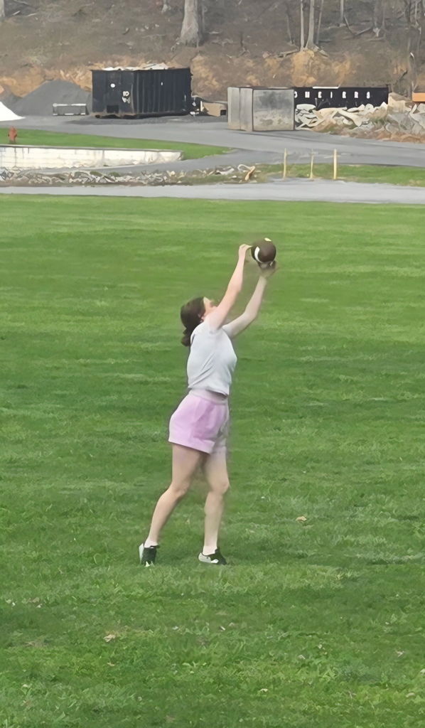 A female student wearing a gray shirt and pink shorts reaches up to catch a football.