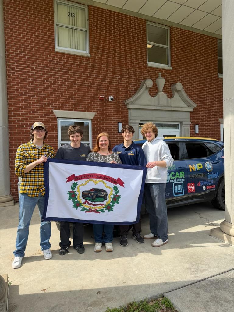 WVU representatives gather around Mrs. Hesse as they all hold up the WV state flag. The blue-colored ECOCar is behind them in the background.