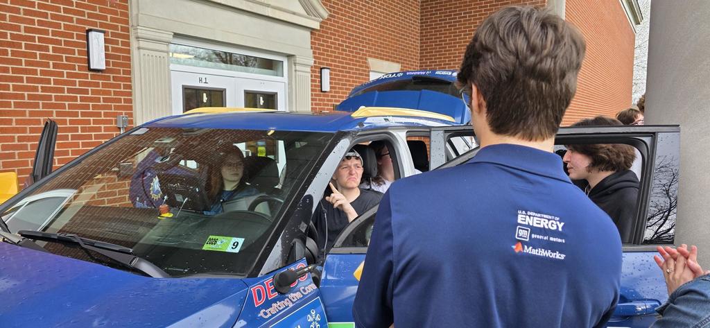 Students gathered around a blue-colored vehicle. There are students inside of the car. The male student in the driver's seat uses ASL to communicate with someone outside of the car.