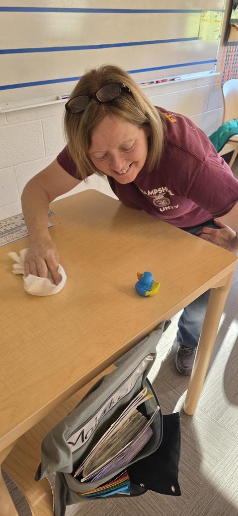 A female custodian wipes off a table while looking down and smiling at her rubber duck.