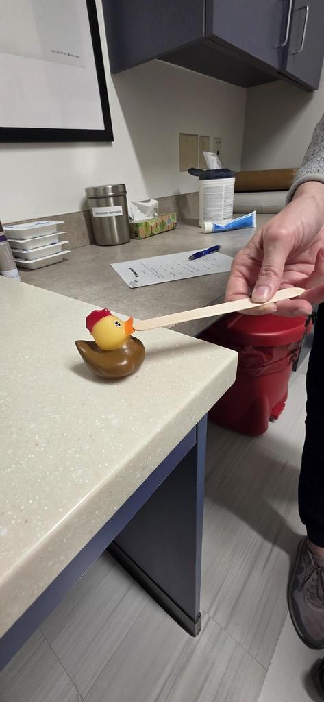 A rubber duck sits on the counter in a doctor's office. Someone is holding a wooden stick to its beak.