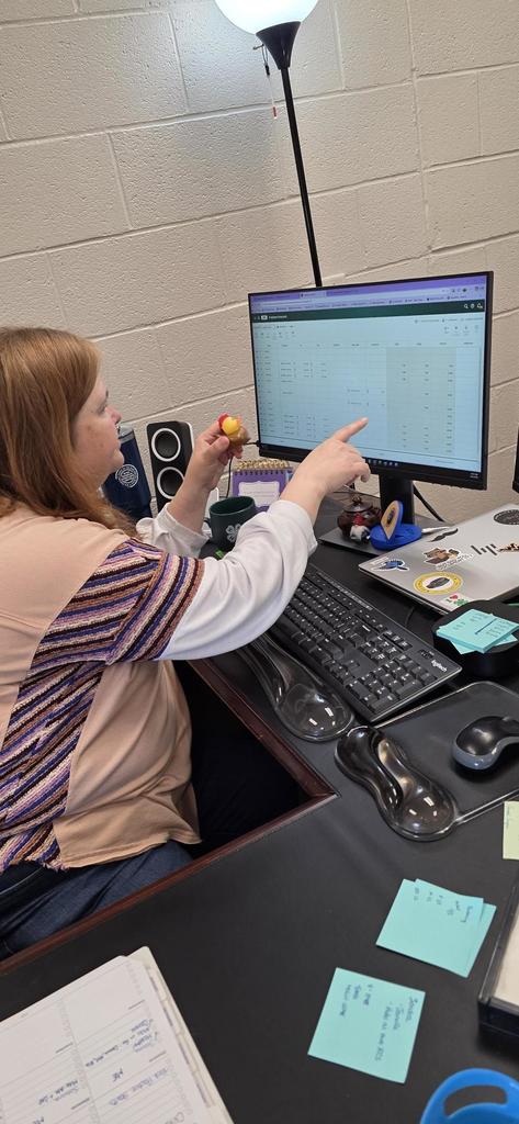 A female, Mrs. Hesse faces a computer screen and points to the screen with her right hand and holds up a rubber duck with her left hand.