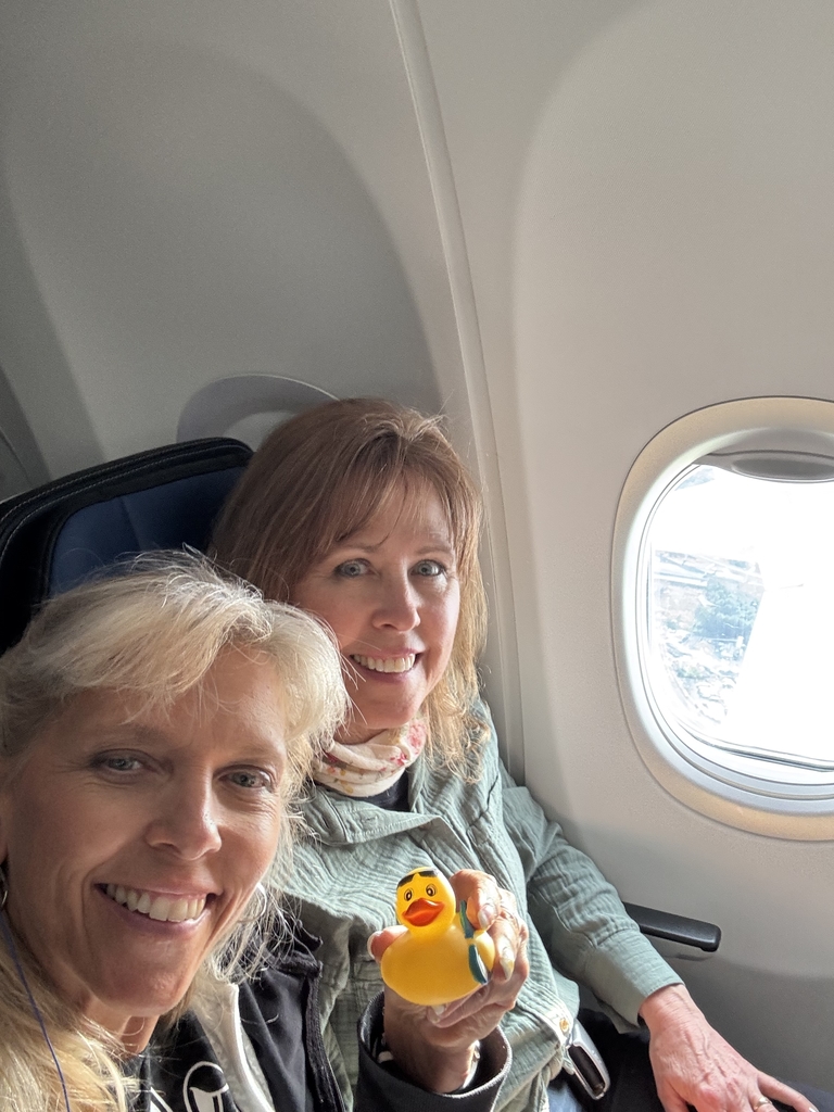 Two females are seated side-by-side in an airplane. The female on the left-hand side holds up a rubber duck.