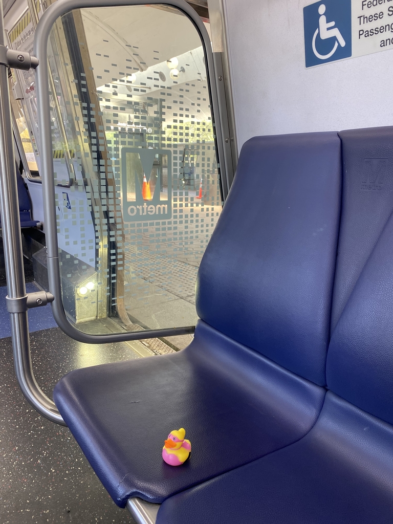 A rubber duck sits on a blue seat inside of a metro train.