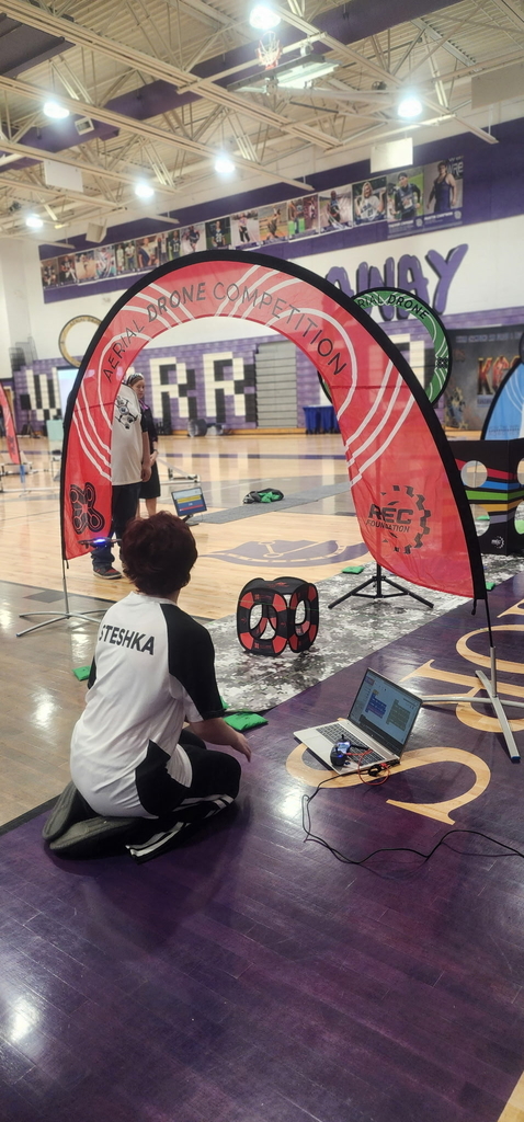 A drone team member sits on the ground with a laptop sitting to the right of them. They have their back away from the camera as they look out towards the drone course.