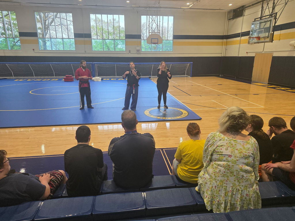 Martial Arts instructors stand on a mat in the gymnasium, along with an ASL interpreter. There are individuals sitting in the bleachers in the foreground.