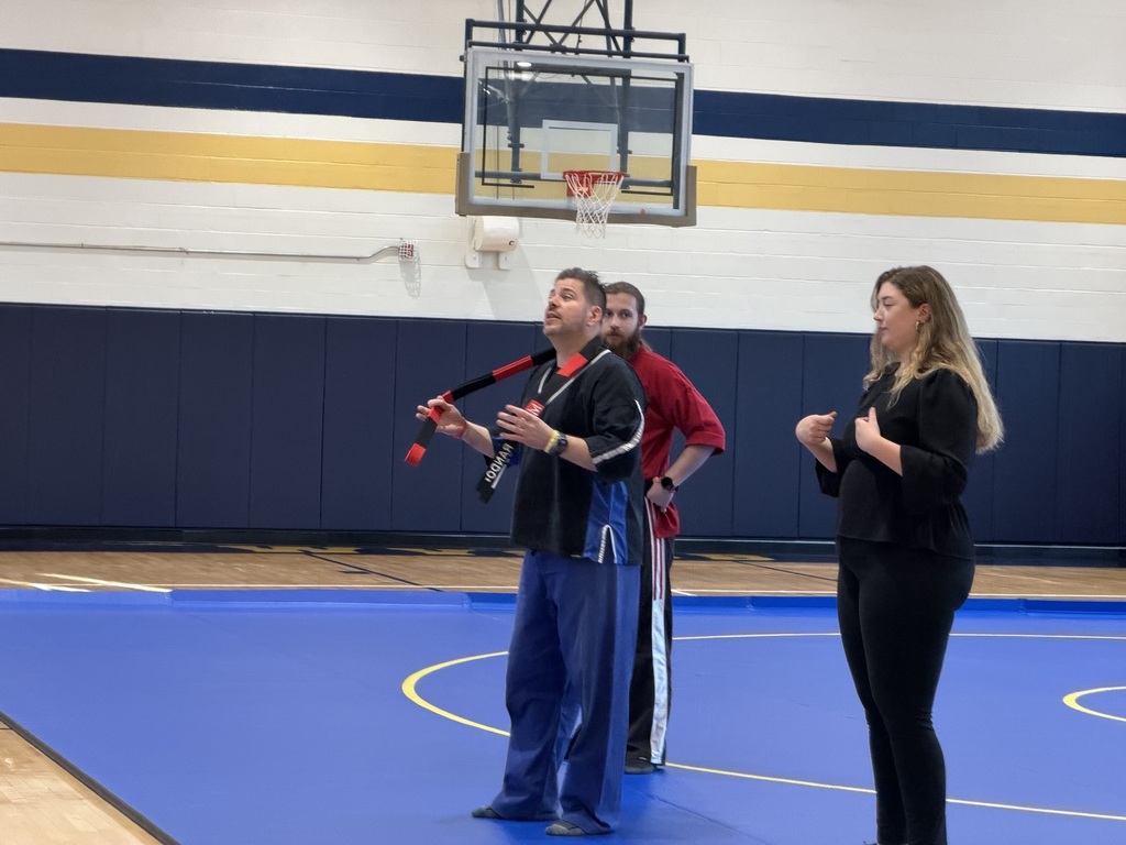 Two male martial arts instructors stand on a blue mat in a gymnasium. There is an ASL interpreter to the right.