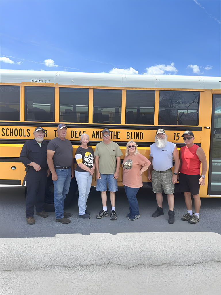 A group of male and female bus drivers stand in front of a yellow bus with "West Virginia Schools for the Deaf and the Blind" in bold, black font.