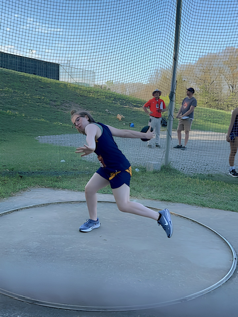 A female track athlete wearing a blue and yellow uniform swings her arm back in preparation to throw a track and field discus.