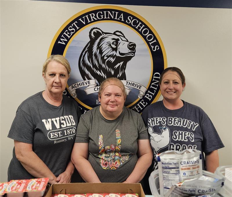 The WVSDB Kitchen Staff. Three female cooks stand side-by-side smiling for the camera in front of a WVSDB Black Bear logo wall decal.