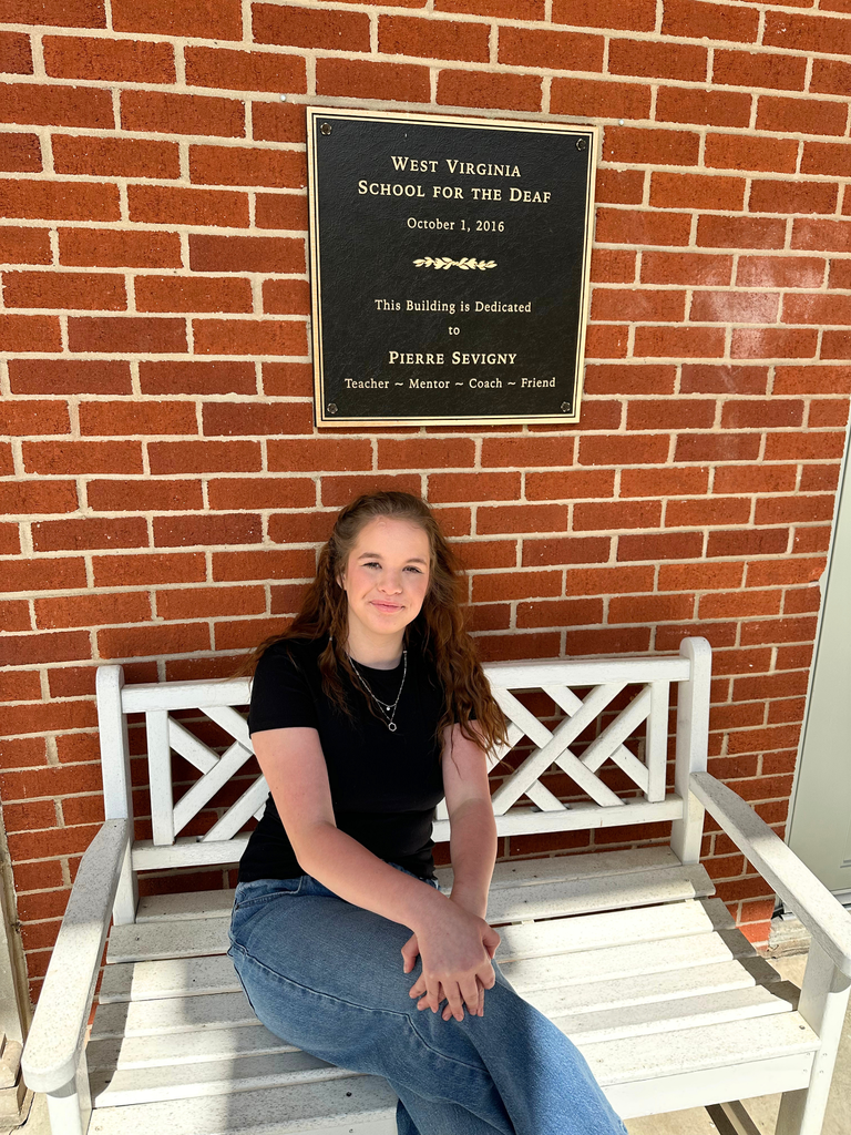 Khloe Bennett, a female student sits on a white bench with her legs crossed and her hands sitting on her lap. There is a brick wall behind her.