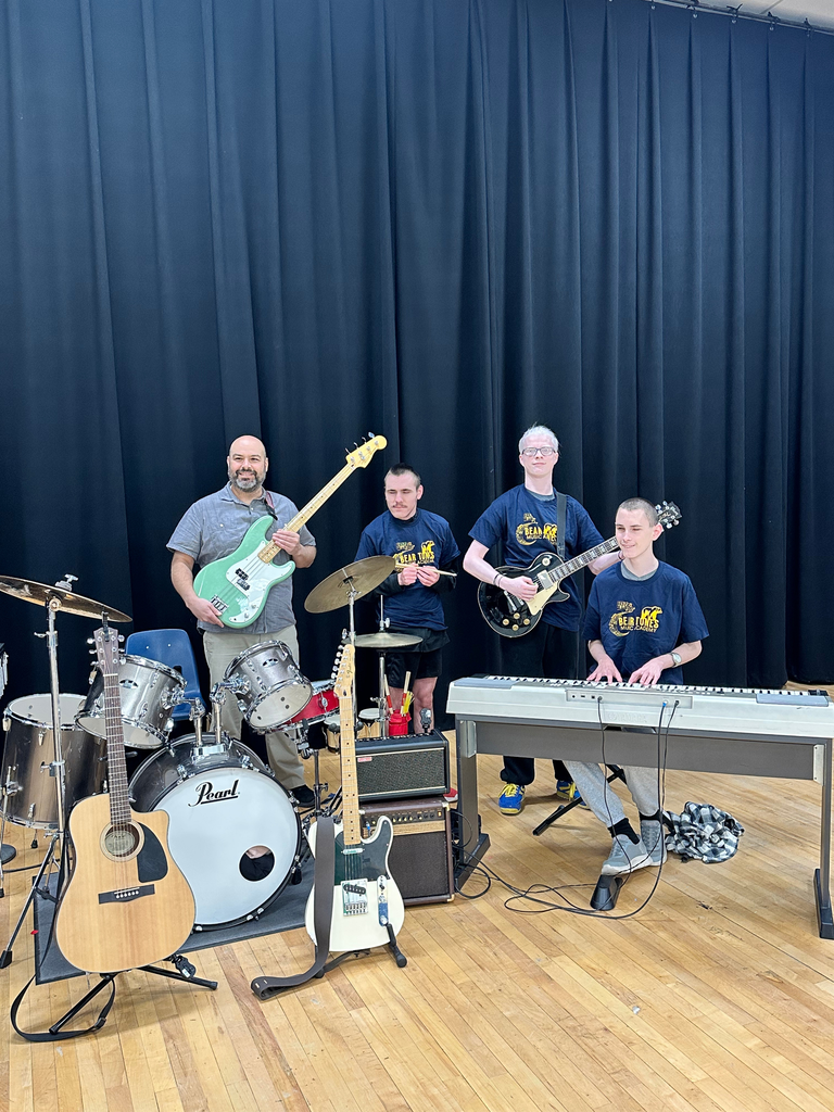 The WVSDB Rock Band pose wit their instruments, alongside Mr. Josh Haza in front of a navy-colored curtain. Instruments include, guitars, drums, and a keyboard.