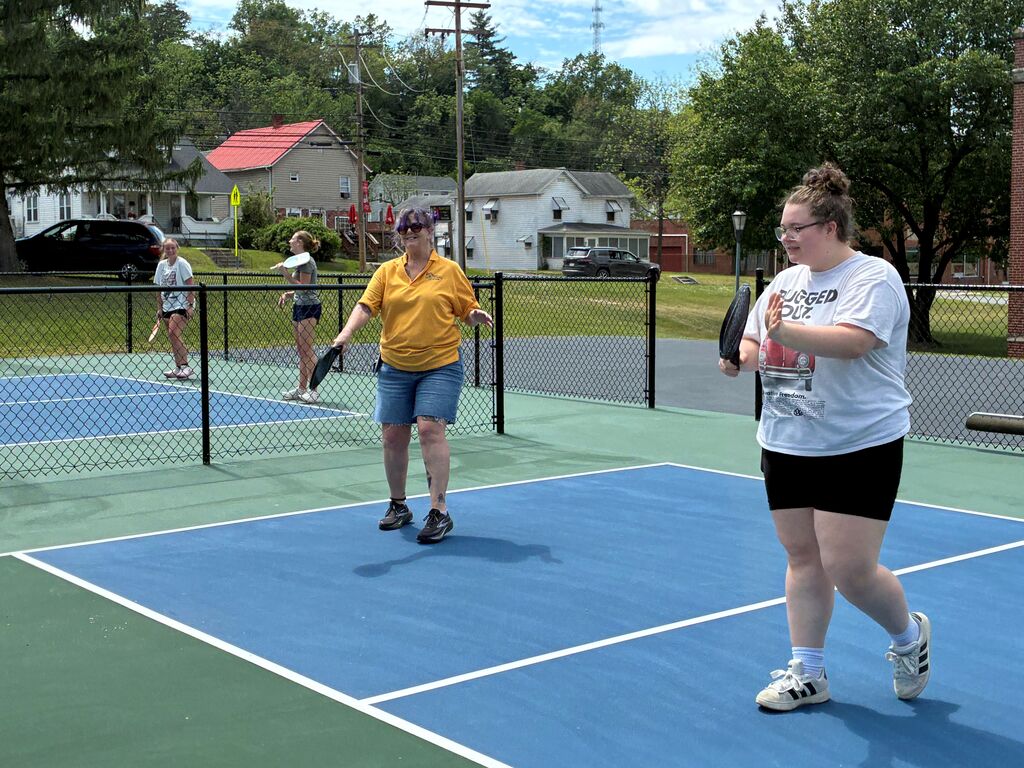 Two females, Sierra and Kim stand on a green and blue-colored court playing a game of pickleball.