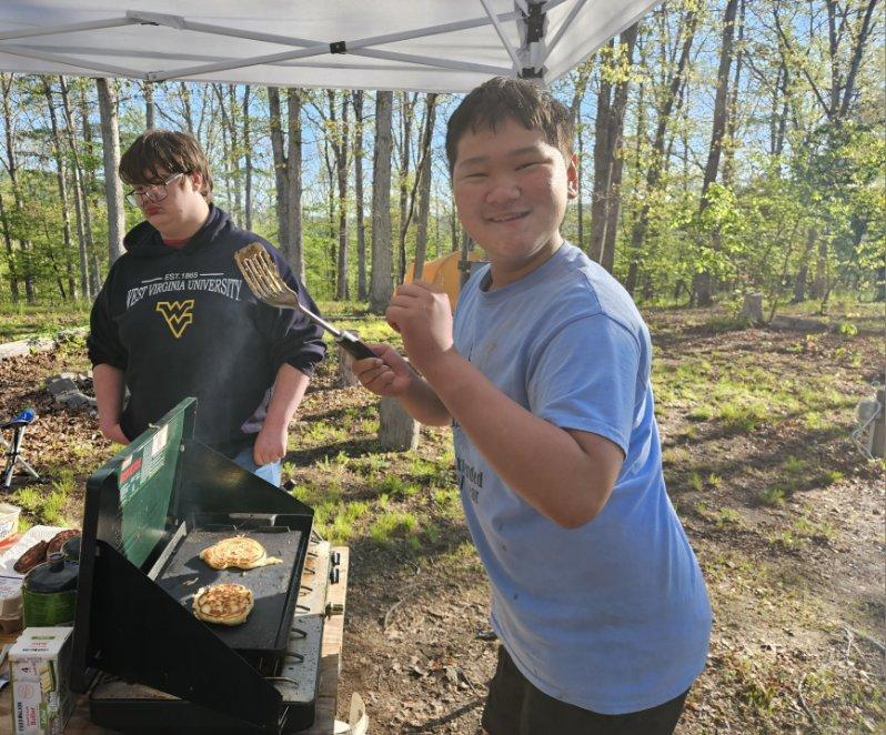 Tristian smiles excitedly as he holds up a spatula. He is preparing pancakes that are cooking on a grill below him.
