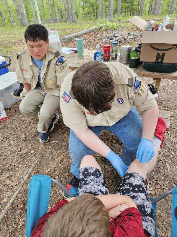 Two Boy Scouts practice doing first-aid on another Scout. One Scout wears blue latex gloves and applies cotton to a wound while another Scout observes.