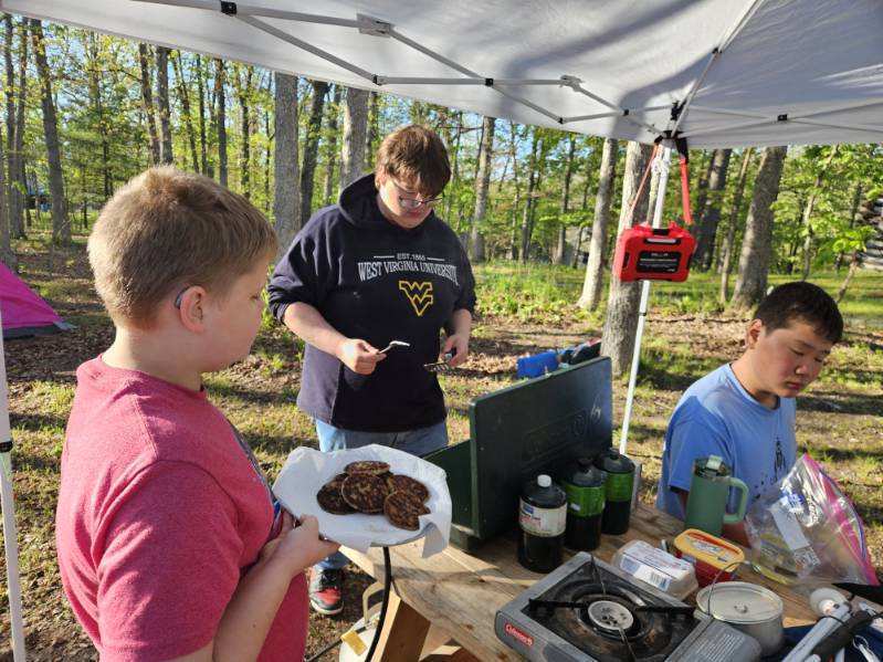 Aiden holds a plate of sausages, while Carson stands behind a grill preparing breakfast items.