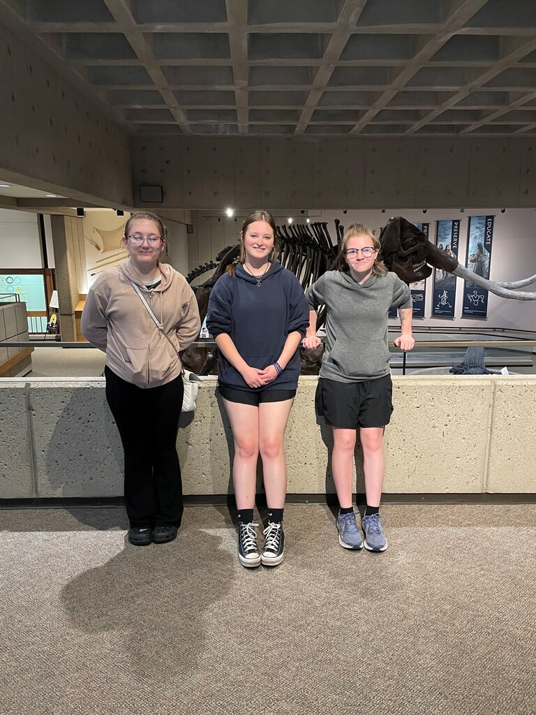 Bray, Madison, and Skylar stand side-by-side with a large fossil in the background while visiting the Archaeological Museum.