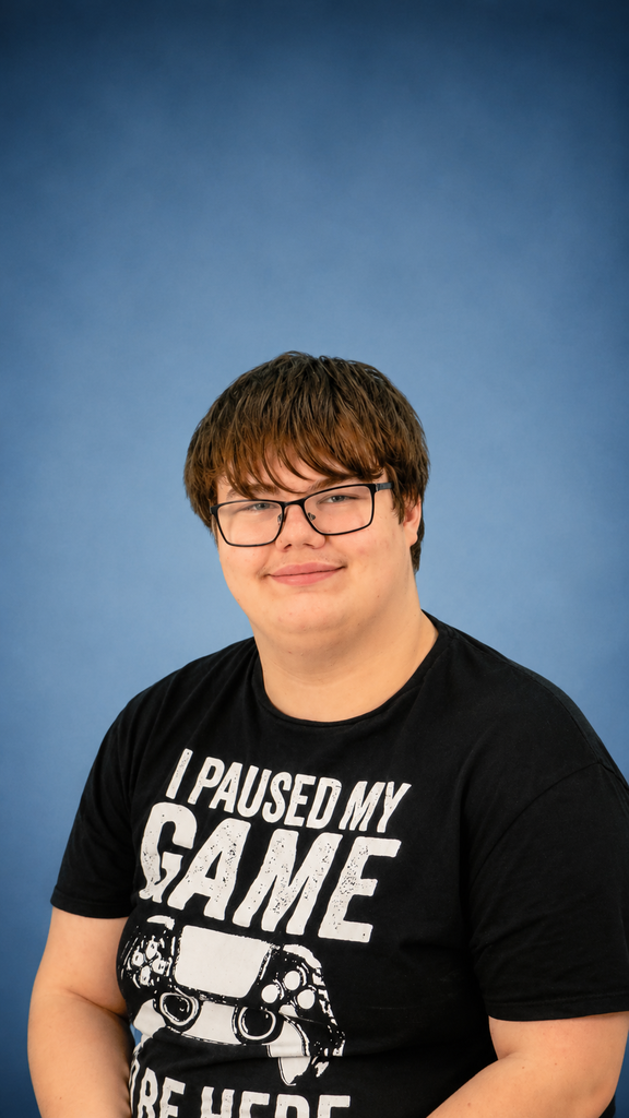 Carson Lawson. Carson smiles for the camera. He is wearing black glasses and a black shirt with white text written on the shirt. He is seated in front of a navy-colored backdrop.