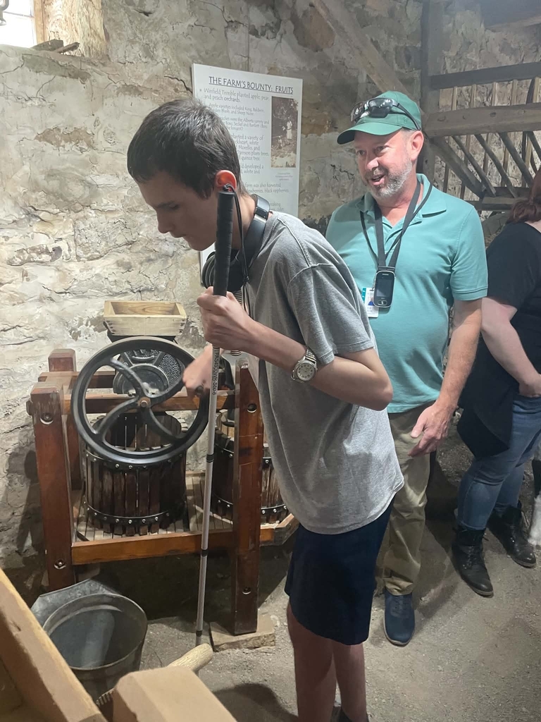A male student, Wyatt, holds his cane in one hand and touches a wheel with his other hand. A male worker smiles and observes Wyatt.