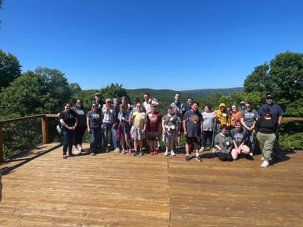 A group photo of students and chaperones standing on a wooden structure with a green mountain range in the background.
