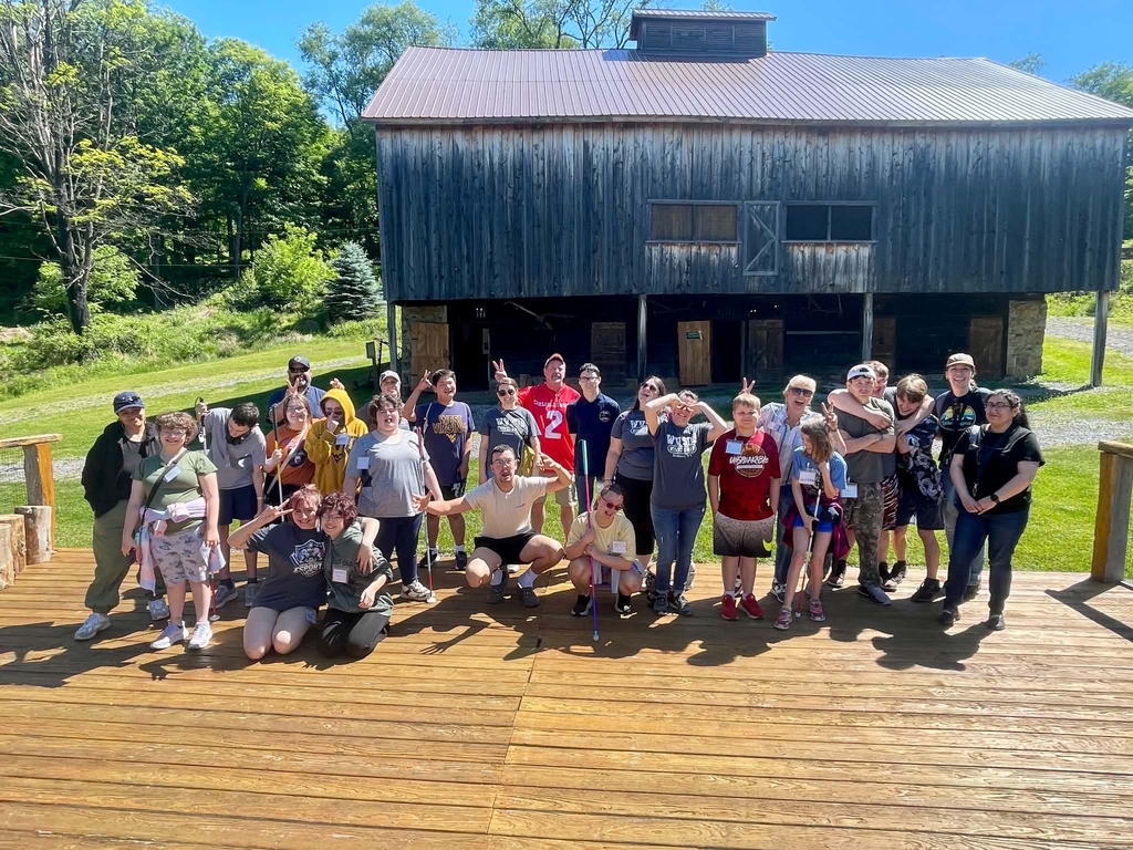 Group photo of students and chaperones pose silly while they stand on a wooden structure with an old, wooden barn in the background.