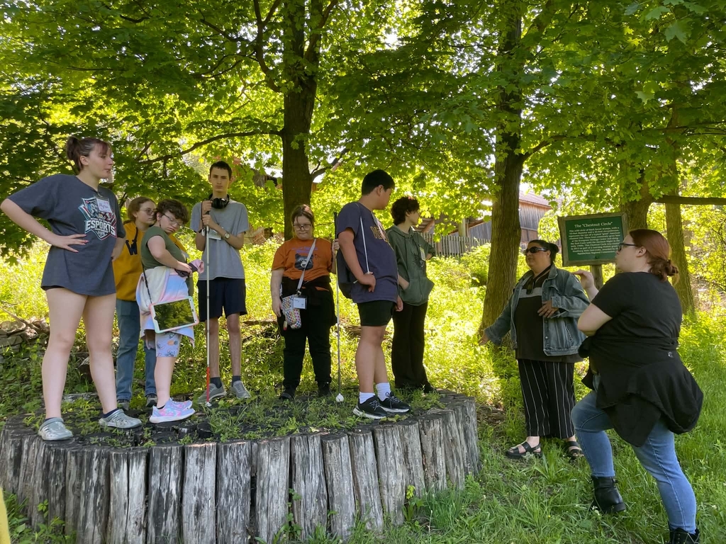 Students standing on a large tree stump while a female speaks and teaches them about the Evergreen Heritage Center.