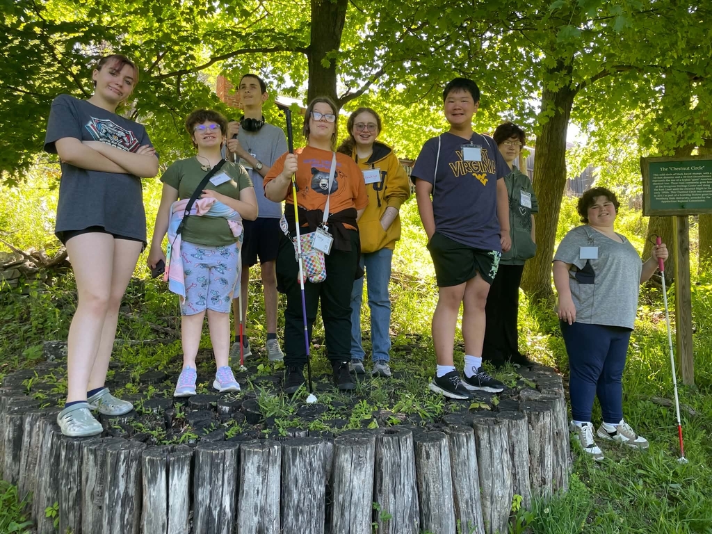 Students are standing on a large tree stump while looking towards the camera and smiling. There are green, earthy trees in the background.