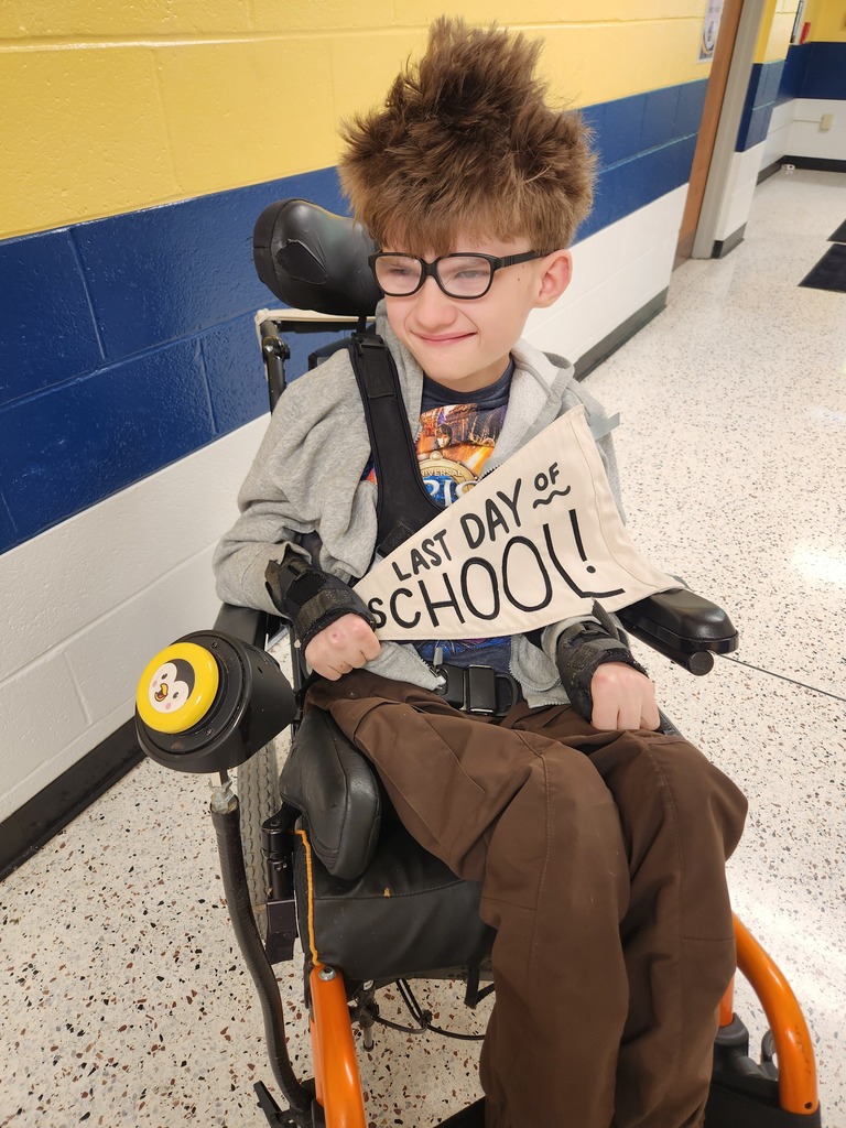 Bryce, a male student seated in a wheelchair smiles and holds up a beige-colored pennant flag with black text "Last Day of School."