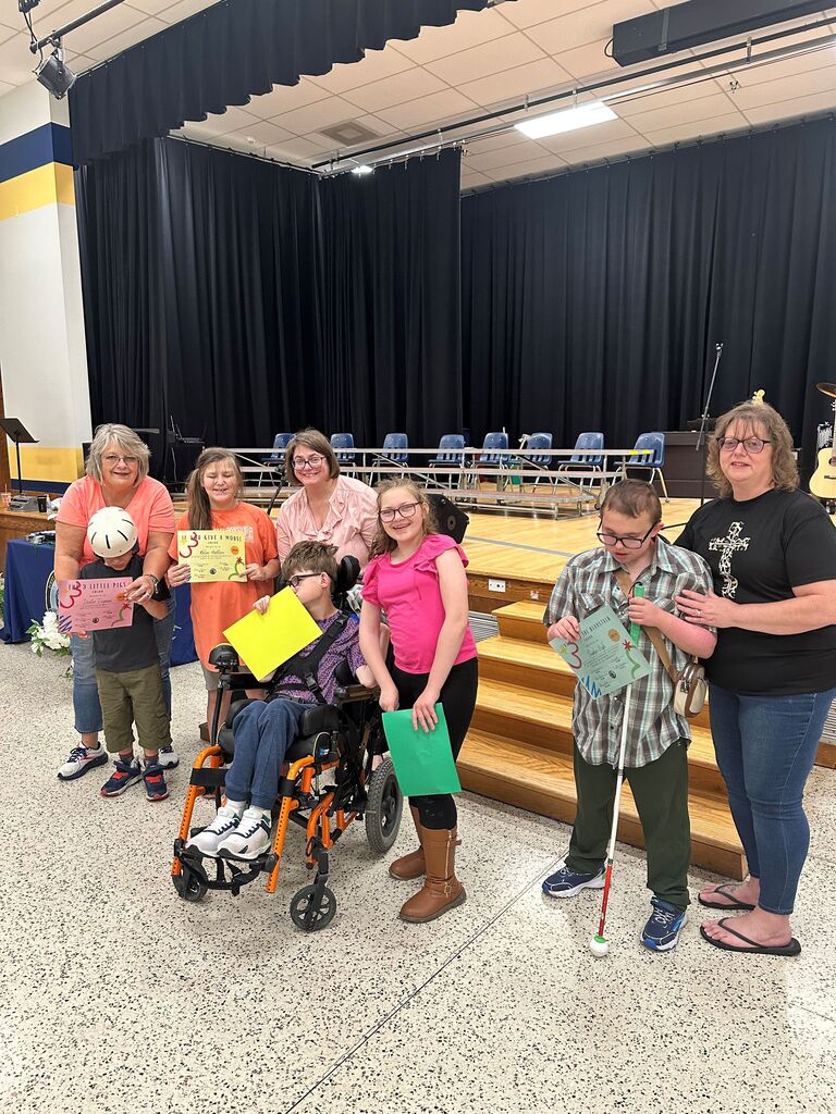Mrs. Park's class stands beside one another holding up their certificates of achievement in front of a stage.
