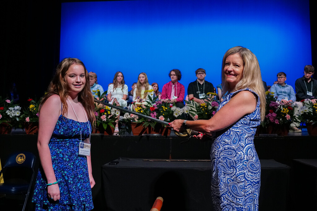 Khloe Bennett, wearing a blue, floral dress smiles for the camera as Superintendent Michele Blatt "knights" her with a sword during the award ceremony. There are seated students in the background.