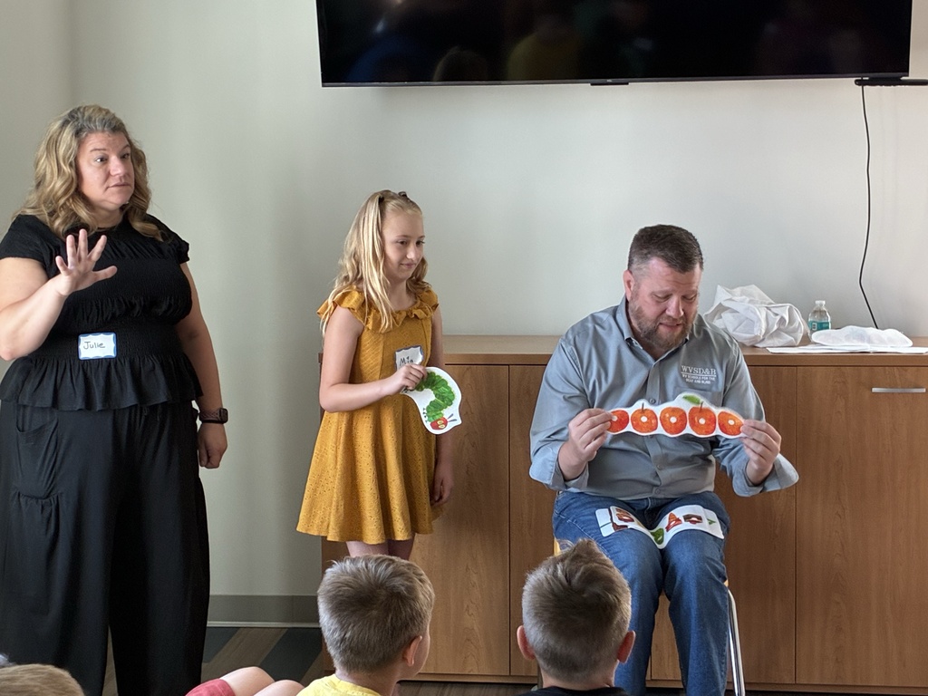 Superintendent Burch holds up paper, red apples while a female attendee wearing a yellow dress holds up a green caterpillar. A female interpreter is to the left of them.