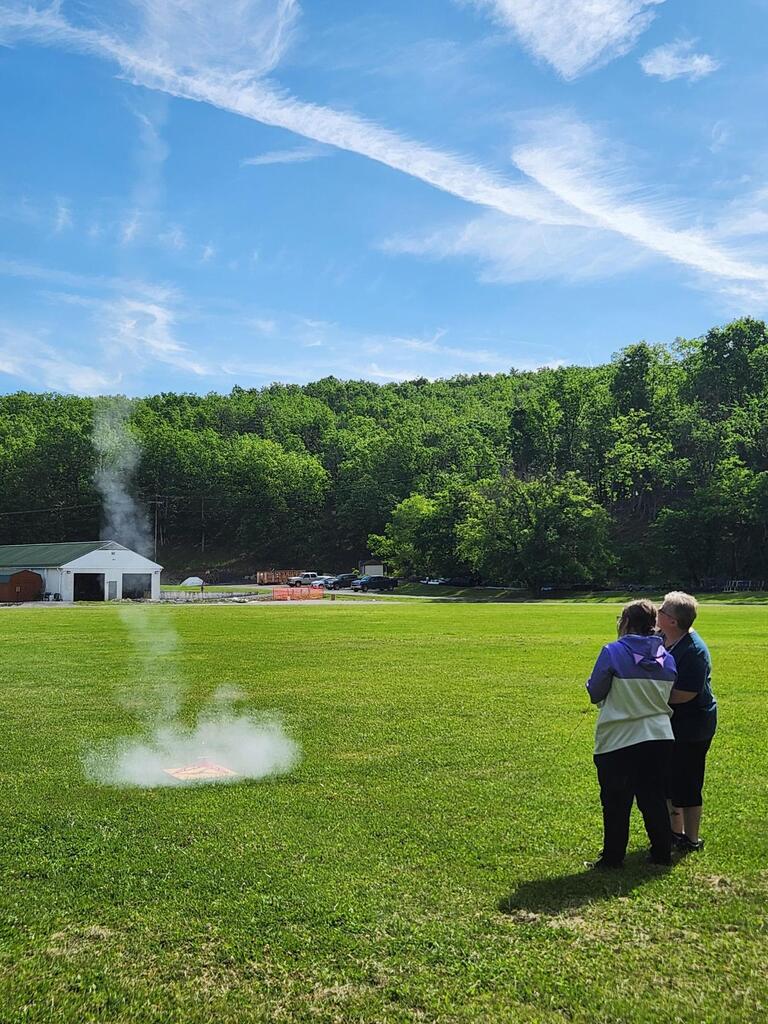 Coach Biddle and Kimmy look up at the sky as they launched Kimmy's rocket. There is smoke coming off of the launchpad after the rocket launched.